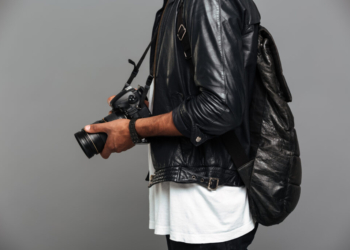 Cropped photo of stylish african man with backpack holding photo camera, isolated on gray background